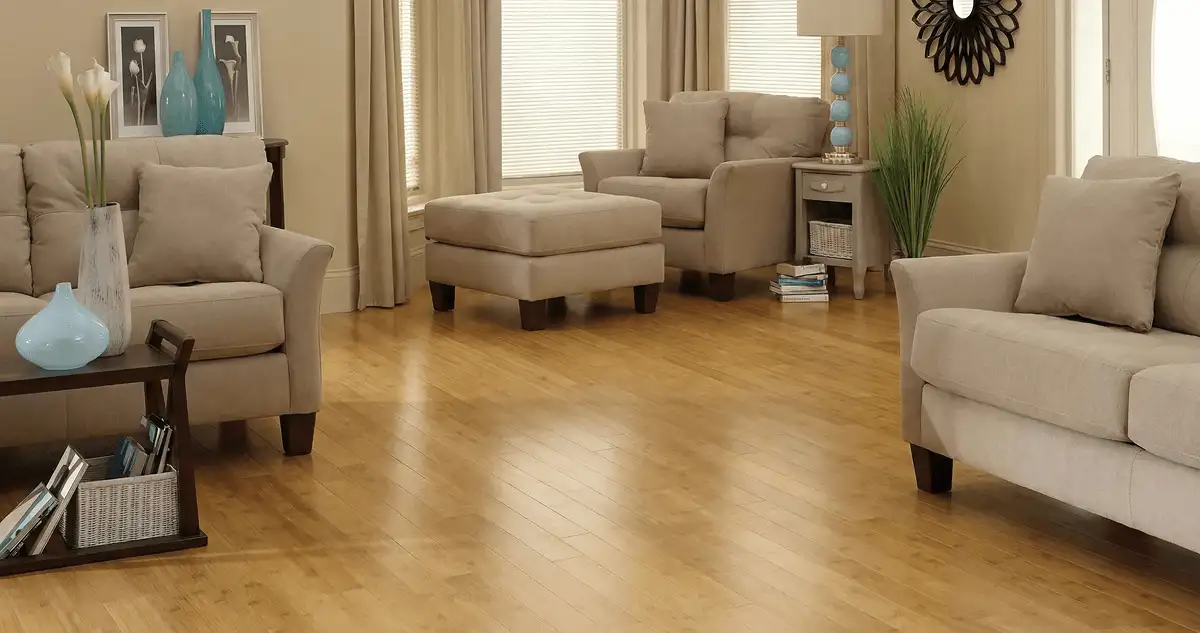 A living room with professionally installed bamboo flooring, beige sofas, a matching ottoman, a side table with a blue lamp, vases, framed pictures, and large windows with blinds and curtains in Boca Raton, FL